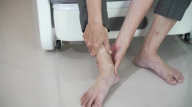 Yaşlı Asyalı adam kanepede otururken ağrıyan ayağına elleriyle masaj yapıyor. Bacak, yaralanma ya da kaval kemiği ve tendonit yüzünden gerginlik. Akut miyozit.