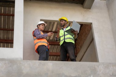 Foreman, mühendis ya da mimar, tamamlanmamış bir binanın inşaat alanındaki gayrimenkul inşaatının detaylarını kontrol etmek için elinde dizüstü bilgisayar ve planlar tutuyor.