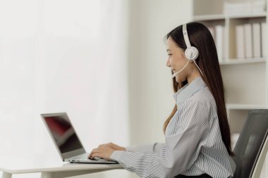 Güzel Asyalı kadın satıcı ya da telsiz kulaklık takan çağrı merkezi operatörü telefonda bir müşteriyle konuşuyor. Bilgisayar görüntülü aramalar yoluyla müşterilere tavsiye ve iletişim sağlayın.