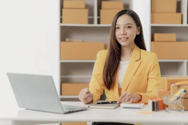 Bağımsız Asyalı kadın KOBİ küçük işletme girişimcisi müşterilere satacak paketler hazırlamak için çevrimiçi siparişleri almak ve gözden geçirmek için dizüstü bilgisayar ve kutular kullanır. KOBİ iş kavramı.