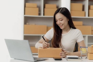 Bağımsız Asyalı kadın KOBİ küçük işletme girişimcisi müşterilere satacak paketler hazırlamak için çevrimiçi siparişleri almak ve gözden geçirmek için dizüstü bilgisayar ve kutular kullanır. KOBİ iş kavramı.