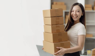 Bağımsız Asyalı kadın KOBİ küçük işletme girişimcisi müşterilere satacak paketler hazırlamak için çevrimiçi siparişleri almak ve gözden geçirmek için dizüstü bilgisayar ve kutular kullanır. KOBİ iş kavramı.