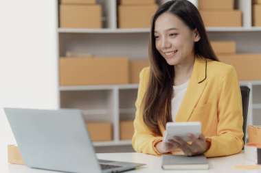 Bağımsız Asyalı kadın KOBİ küçük işletme girişimcisi müşterilere satacak paketler hazırlamak için çevrimiçi siparişleri almak ve gözden geçirmek için dizüstü bilgisayar ve kutular kullanır. KOBİ iş kavramı.