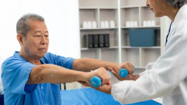 Yaşlı Asyalı bir adam, kıdemli bir kadın terapist hemşirenin desteğiyle fizik tedavi görüyor. Yaşlı adam evde bakım ve sağlık hizmetleri konseptinde hasta için elastik bant kullanıyor..