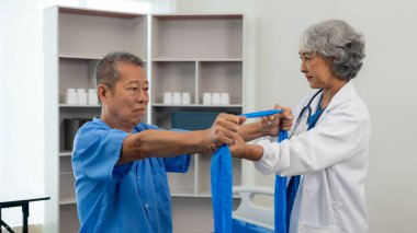 Yaşlı Asyalı bir adam, kıdemli bir kadın terapist hemşirenin desteğiyle fizik tedavi görüyor. Yaşlı adam evde bakım ve sağlık hizmetleri konseptinde hasta için elastik bant kullanıyor..
