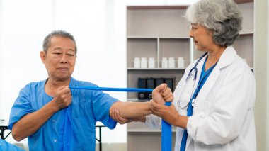 Yaşlı Asyalı bir adam, kıdemli bir kadın terapist hemşirenin desteğiyle fizik tedavi görüyor. Yaşlı adam evde bakım ve sağlık hizmetleri konseptinde hasta için elastik bant kullanıyor..