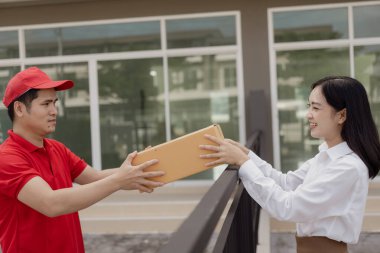 Kırmızı elbiseli genç kurye karton bir paket kutusu tutuyor ve kadınlar paketleri evden, online alışveriş ve mal teslimatından almak için cep telefonuyla para transfer ediyorlar..