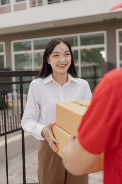 Kırmızı elbiseli genç kurye karton bir paket kutusu tutuyor ve kadınlar paketleri evden, online alışveriş ve mal teslimatından almak için cep telefonuyla para transfer ediyorlar..