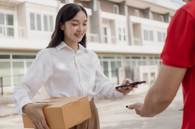 Kırmızı elbiseli genç kurye karton bir paket kutusu tutuyor ve kadınlar paketleri evden, online alışveriş ve mal teslimatından almak için cep telefonuyla para transfer ediyorlar..