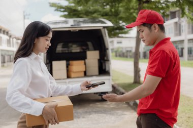 Kırmızı elbiseli genç kurye karton bir paket kutusu tutuyor ve kadınlar paketleri evden, online alışveriş ve mal teslimatından almak için cep telefonuyla para transfer ediyorlar..