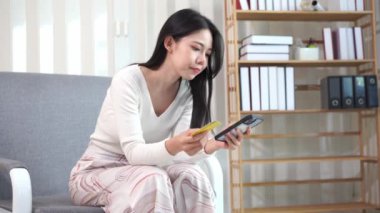 Genç Asyalı bir kadın, plastik bir kredi kartıyla ev telefonunun rahatlığıyla online alışveriş yapıyor. Evde para transferi, oturma odasında kanepede oturan güzel bir kadın.