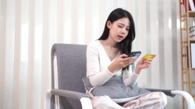 Genç Asyalı bir kadın, plastik bir kredi kartıyla ev telefonunun rahatlığıyla online alışveriş yapıyor. Evde para transferi, oturma odasında kanepede oturan güzel bir kadın.