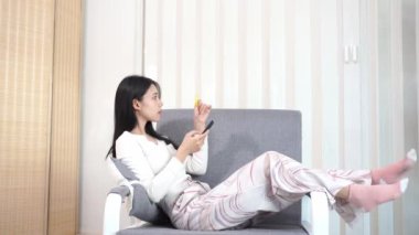 Genç Asyalı bir kadın, plastik bir kredi kartıyla ev telefonunun rahatlığıyla online alışveriş yapıyor. Evde para transferi, oturma odasında kanepede oturan güzel bir kadın.