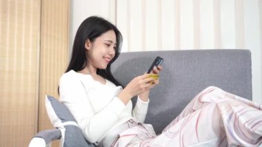 Genç Asyalı bir kadın, plastik bir kredi kartıyla ev telefonunun rahatlığıyla online alışveriş yapıyor. Evde para transferi, oturma odasında kanepede oturan güzel bir kadın.