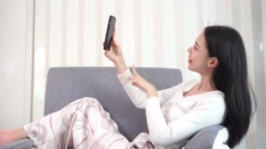 Evdeki koltukta oturan genç Asyalı kadın cep telefonuyla mesaj atıyor ve arkadaşlarıyla telefonda konuşuyor. Ve arkadaşlarınla video görüşmelerinin tadını çıkar. rahatlama konsepti