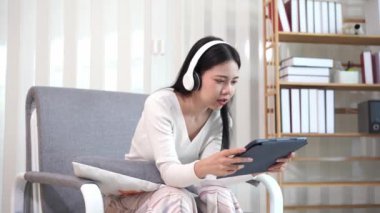 Asyalı kız tabletle mutlu bir şekilde oynuyor, rahat bir koltukta oturuyor, online alışveriş yapıyor sosyal medya uygulamalarını kullanarak internette sörf yapıyor ya da modern teknoloji cihazlarıyla oyunlar oynuyor..