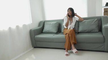 Rahat gülümseyen genç bir kadın koltukta oturuyor cep telefonu teknolojisini kullanıyor gezin ve mobil uygulamadan online e-ticaret alışverişinin tadını çıkarın ve gömülü film ve müzik izleyin..