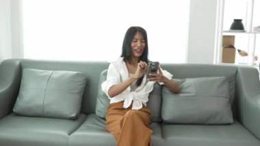 Rahat gülümseyen genç bir kadın koltukta oturuyor cep telefonu teknolojisini kullanıyor gezin ve mobil uygulamadan online e-ticaret alışverişinin tadını çıkarın ve gömülü film ve müzik izleyin..