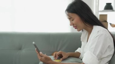 Rahat gülümseyen genç bir kadın koltukta oturuyor cep telefonu teknolojisini kullanıyor gezin ve mobil uygulamadan online e-ticaret alışverişinin tadını çıkarın ve gömülü film ve müzik izleyin..