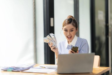Asyalı iş kadını elinde para tutuyor, iş başarısından memnun, masaya oturmuş dolar tutan mutlu Asyalı kadın ve takım elbise içinde gülümsüyor, başarı kavramı.