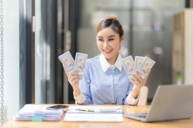 Asyalı iş kadını elinde para tutuyor, iş başarısından memnun, masaya oturmuş dolar tutan mutlu Asyalı kadın ve takım elbise içinde gülümsüyor, başarı kavramı.