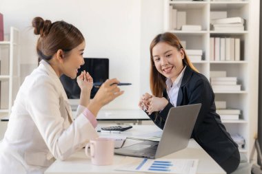 İki Asyalı iş kadını birlikte çalışıyor, yeni bir iş projesiyle tanışıyor ve tartışıyorlar. Yaratıcı ekip, pazarlama planı laptop ve masadaki belgeleri inceliyor.