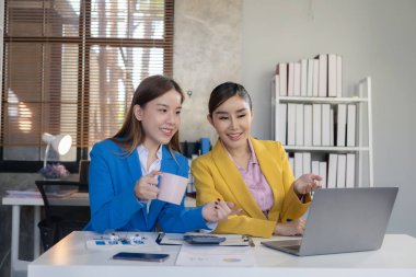 İki Asyalı iş kadını birlikte çalışıyor, yeni bir iş projesiyle tanışıyor ve tartışıyorlar. Yaratıcı ekip, pazarlama planı laptop ve masadaki belgeleri inceliyor.