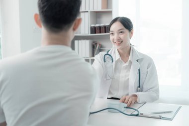 Asyalı kadın hemşire konuşuyor ve genç erkek hasta semptomları hakkında soru soruyor tansiyon monitörü kullanarak muayene sırasında kan basıncı seviyesini ölçüyor.