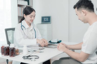 Asyalı kadın hemşire konuşuyor ve genç erkek hasta semptomları hakkında soru soruyor tansiyon monitörü kullanarak muayene sırasında kan basıncı seviyesini ölçüyor.