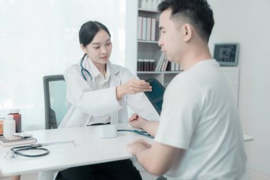 Asyalı kadın hemşire konuşuyor ve genç erkek hasta semptomları hakkında soru soruyor tansiyon monitörü kullanarak muayene sırasında kan basıncı seviyesini ölçüyor.