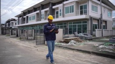Genç profesyonel mühendis bir konut projesini inceliyor, elinde tablet var, binaya bakıyor, başlık takıyor. 4K video, yavaş çekim.