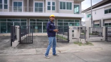 Genç profesyonel mühendis bir konut projesini inceliyor, elinde tablet var, binaya bakıyor, başlık takıyor. 4K video, yavaş çekim.