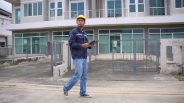 Genç profesyonel mühendis bir konut projesini inceliyor, elinde tablet var, binaya bakıyor, başlık takıyor. 4K video, yavaş çekim.