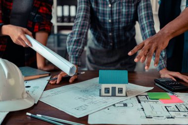 Mimarlar ve mühendisler, inşaat planlarını tartışmak üzere bina içindeki mimari çizimlerin ayrıntılı planlarına ve etrafa saçılmış mimar şapkaları ve mimari belgelere işaret ediyorlar. Yakın plan..