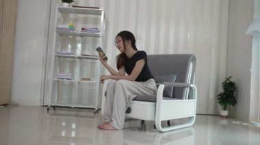 Güzel mutlu Asyalı kadın evdeki oturma odasında koltukta oturuyor, cep telefonunu elinde tutuyor, konuşuyor ve sosyal medyayı kullanıyor. Kanepede internette sörf yap..