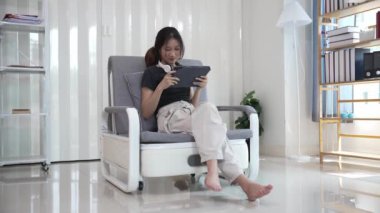 Genç Asyalı kadın elinde dijital tabletle kanepede oturmuş internette sörf yapıyor, sosyal medya uygulaması kullanarak online alışveriş yapıyor..
