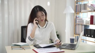 Asyalı bir kadın dizüstü bilgisayar kullanıyor ve hesap makinesi dokümanlarıyla iş raporları yatırım planlarını analiz etmek için merkez büroda çalışıyor.