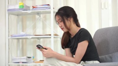 Güzel mutlu Asyalı kadın evdeki oturma odasında koltukta oturuyor, cep telefonunu elinde tutuyor, konuşuyor ve sosyal medyayı kullanıyor. Kanepede internette sörf yap..