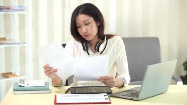 Asyalı bir kadın dizüstü bilgisayar kullanıyor ve hesap makinesi dokümanlarıyla iş raporları yatırım planlarını analiz etmek için merkez büroda çalışıyor.