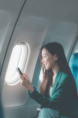 Uçaktaki genç Asyalı kadın, uçak kabininde kıtalararası uçuşlar sırasında internetten film izlerken akıllı telefon kullanıyor. Makinedeki kablosuz bağlantıyı kullanmaktan mutlu.
