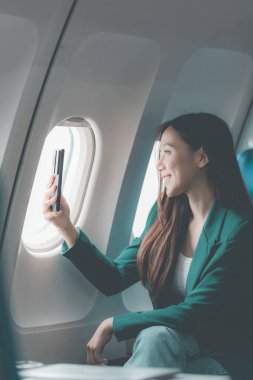 Uçaktaki genç Asyalı kadın, uçak kabininde kıtalararası uçuşlar sırasında internetten film izlerken akıllı telefon kullanıyor. Makinedeki kablosuz bağlantıyı kullanmaktan mutlu.