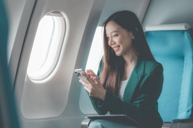 Uçaktaki genç Asyalı kadın, uçak kabininde kıtalararası uçuşlar sırasında internetten film izlerken akıllı telefon kullanıyor. Makinedeki kablosuz bağlantıyı kullanmaktan mutlu.
