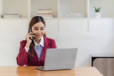 Asyalı kadın ofiste dizüstü bilgisayarla masada otururken akıllı telefon kullanıyor. Belge grafikleri üzerine notlar yazıyor. Finansal büyüme istatistiklerine sahip kadın mali muhasebeci