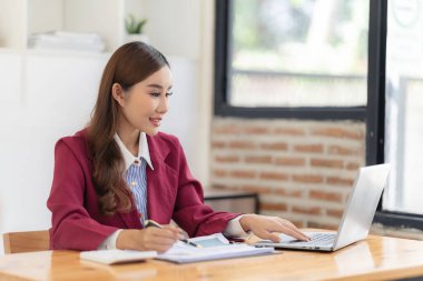 Hesap makinesiyle çalışan gülümseyen Asyalı muhasebeci masadaki belgelerle çevrili dizüstü bilgisayardaki finansal belgeleri analiz ediyor.