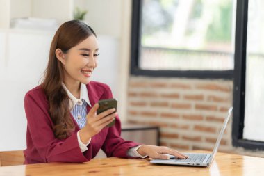 Asyalı kadın ofiste dizüstü bilgisayarla masada otururken akıllı telefon kullanıyor. Belge grafikleri üzerine notlar yazıyor. Finansal büyüme istatistiklerine sahip kadın mali muhasebeci