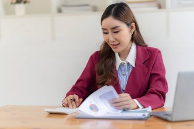 Hesap makinesiyle çalışan gülümseyen Asyalı muhasebeci masadaki belgelerle çevrili dizüstü bilgisayardaki finansal belgeleri analiz ediyor.