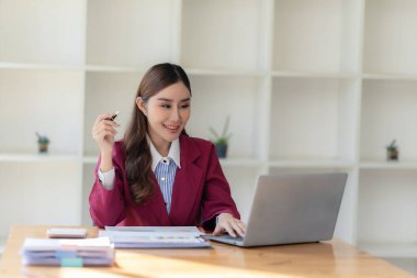 Hesap makinesiyle çalışan gülümseyen Asyalı muhasebeci masadaki belgelerle çevrili dizüstü bilgisayardaki finansal belgeleri analiz ediyor.