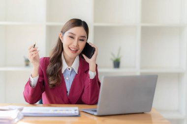 Asyalı kadın ofiste dizüstü bilgisayarla masada otururken akıllı telefon kullanıyor. Belge grafikleri üzerine notlar yazıyor. Finansal büyüme istatistiklerine sahip kadın mali muhasebeci