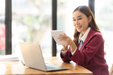 Hesap makinesiyle çalışan gülümseyen Asyalı muhasebeci masadaki belgelerle çevrili dizüstü bilgisayardaki finansal belgeleri analiz ediyor.
