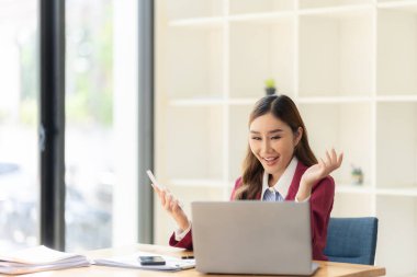 Hesap makinesiyle çalışan gülümseyen Asyalı muhasebeci masadaki belgelerle çevrili dizüstü bilgisayardaki finansal belgeleri analiz ediyor.